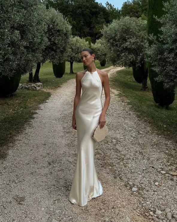 The Jeany Silk Dress delights with adjustable straps and a chic halter neckline, perfectly complemented by elegant back buttons and bias stitching that beautifully drapes your silhouette. Designed to flirt with the floor when paired with heels, it offers a stunningly elongated look—just the right excuse for a little hemming fun! The playful ties at the back ensure a fit as unique as you are, combining charm and comfort for those unforgettable special moments.