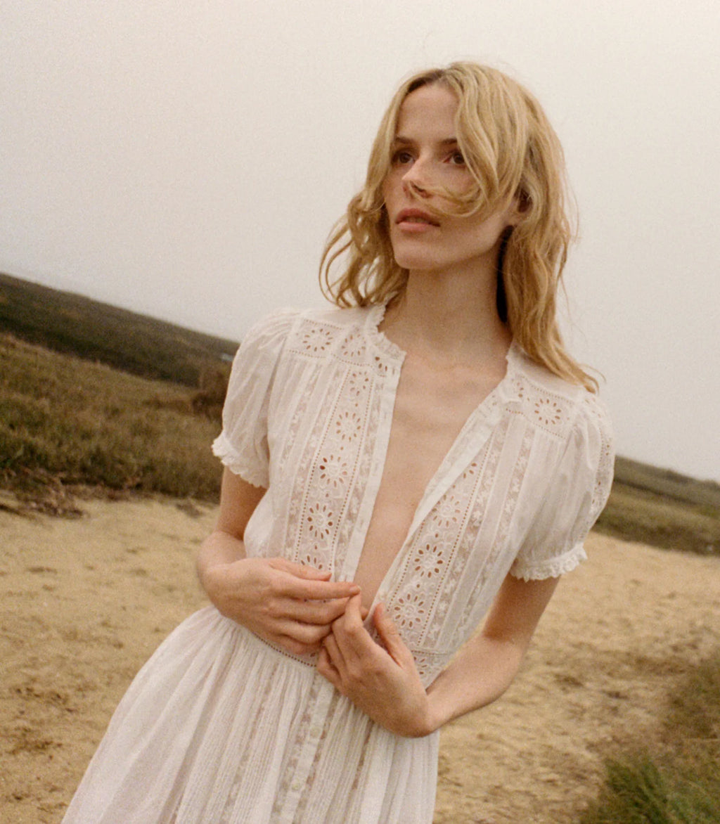 Experience effortless elegance with the Windsor Dress by Doen, crafted from the softest organic cotton voile for ultimate comfort. Inspired by our beloved Leigh Top, it features charming short puffed sleeves and intricate alternating panels of lace, eyelet embroidery, and ladder trim. A delicate row of starburst buttons adorns the front, while a fitted waist flows into an ankle-length skirt with a graceful ruffle, combining timeless style with a flattering silhouette. 